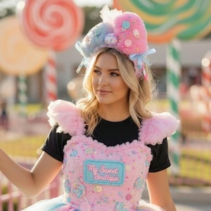 Puede incluir: Un atuendo rosa y azul con temática de dulces y una diadema a juego. El vestido presenta un corpiño con el texto "My Sweet Boutique" y está adornado con adornos en forma de caramelo. La diadema tiene detalles de caramelos de felpa.