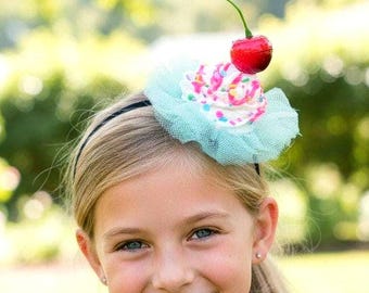 May include: A headband featuring a cupcake decoration with white frosting, colorful sprinkles, and a bright red cherry on top. The cupcake sits on a ruffled light blue tulle base, attached to a thin black band that rests on the head.