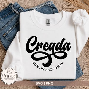 May include: A white long-sleeve sweatshirt featuring the black text Creada in a bold script font, with the text CON UN PROPOSITO printed in a smaller sans-serif font underneath. The sweatshirt has a crew neckline and long sleeves with ribbed cuffs.
