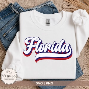 May include: A white long-sleeve crewneck sweatshirt featuring the word Florida in a bold, cursive script font. The text is layered with a white top, a dark blue middle, and a red bottom outline, creating a three-dimensional effect on the fabric.