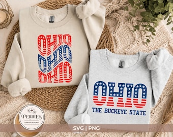May include: A beige crewneck sweatshirt featuring a wavy, red and blue American flag-patterned Ohio graphic, alongside a light grey crewneck sweatshirt displaying the text OHIO THE BUCKEYE STATE in a blue and red American flag-inspired font.