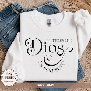 Op de afbeelding: Een wit sweatshirt met lange mouwen en ronde hals met zwarte tekst die EL TIEMPO DE Dios ES PERFECTO leest in een mix van elegante schreefletters en decoratieve scriptlettertypes. Het kledingstuk is opgevouwen en getoond met een blauwe jeans.