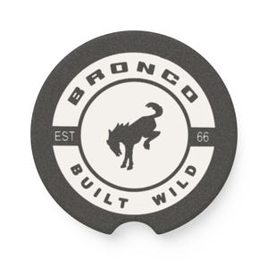 May include: A circular dark gray car coaster featuring a white center circle with a black bucking horse silhouette. The text BRONCO is arched at the top, BUILT WILD is arched at the bottom, and EST 66 is printed on the sides of the center circle.