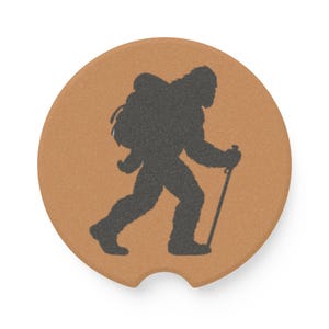 May include: A round, tan-colored cork car coaster featuring a dark gray silhouette of a creature resembling Bigfoot, depicted walking with a backpack and a hiking pole. The coaster has a small semi-circular notch cut out from the bottom edge.