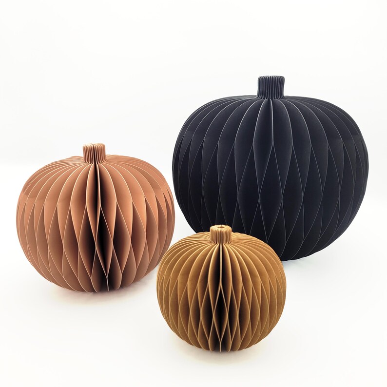 May include: Three decorative paper spheres in varying sizes and colors. The largest is black, the medium is a copper-brown, and the smallest is a golden-brown. Each sphere has a ribbed texture and a small, cylindrical top, suggesting a vase or decorative object.