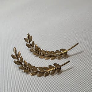 May include: Two gold-colored leaf ear cuffs. Each cuff features a curved design of detailed leaves, creating a botanical aesthetic. The ear cuffs are made of a metallic material and are displayed on a white surface.
