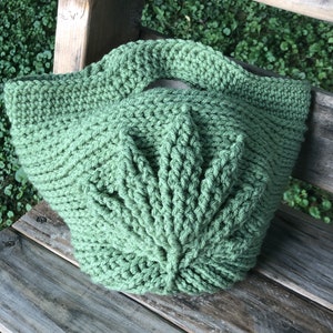 May include: A green crocheted tote bag with a unique leaf-shaped design. The bag has a single handle and is made with a thick yarn.