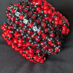 May include: A multi-layered cuff bracelet constructed from small, round, opaque red and black plastic pony beads. The beads are woven together in a dense, textured pattern. Three white square letter beads spell out the letters M, C, and R.