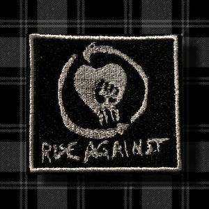 Puede incluir: Parche bordado en blanco y negro con el logotipo de Rise Against. El logotipo presenta una calavera dentro de un corazón con un círculo alrededor. El texto "Rise Against" está debajo del logotipo.