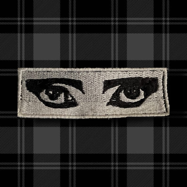 May include: A white embroidered patch with two black eyes. The eyes are large and have a dramatic look.
