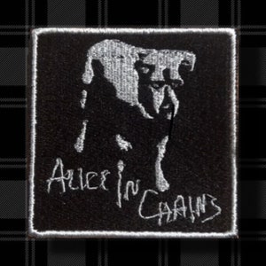 May include: Black square patch featuring a white illustration of a dog-like creature. The text "Alice in Chains" is embroidered in white cursive font below the illustration. The patch is set against a black and gray plaid background.