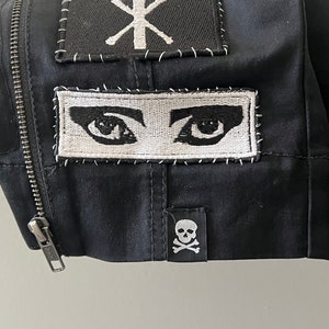 May include: Black jacket with three embroidered patches. The top patch is black with white stitching and says "Tribal Death" with a cross symbol. The middle patch is white with black stitching and shows two eyes. The bottom patch is black with white stitching and shows a skull and crossbones.