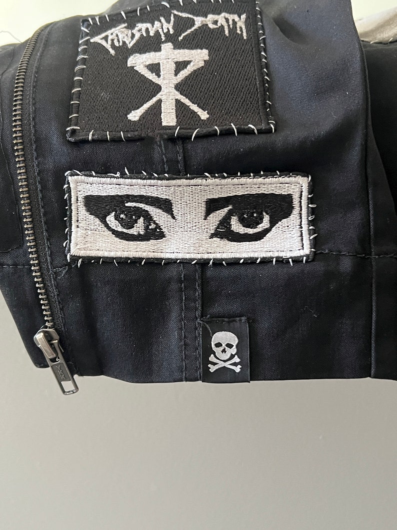 May include: Black jacket with three embroidered patches. The top patch is black with white stitching and says "Tribal Death" with a cross symbol. The middle patch is white with black stitching and shows two eyes. The bottom patch is black with white stitching and shows a skull and crossbones.