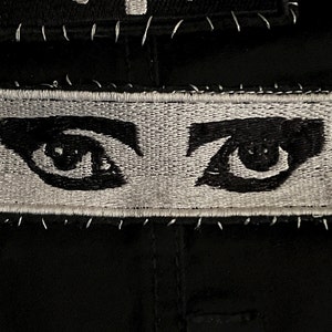 May include: A black and white embroidered patch with two eyes. The eyes are black with white pupils and are surrounded by white eyelashes. The patch is sewn onto a black fabric.