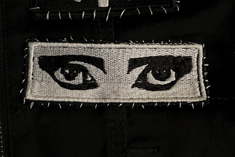 May include: A black and white embroidered patch with two eyes. The eyes are black with white pupils and are surrounded by white eyelashes. The patch is sewn onto a black fabric.