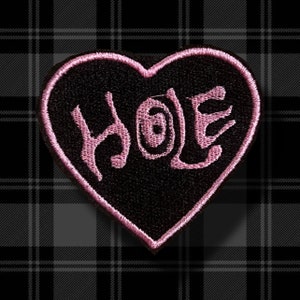 May include: Pink heart-shaped patch with the word "HOLE" written in pink on a black background.