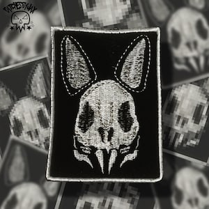 May include: White embroidered bunny skull patch on a black background. The patch has a stitched border.