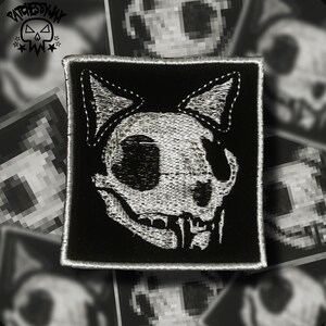 May include: Black and white embroidered patch of a cat skull with a stitched outline. The patch is square and has a black background.