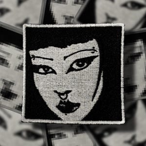 May include: Square black and white embroidered patch featuring a stylized face with dark eyes, a nose ring, and full lips. The patch has a black border and is likely for clothing or accessories.