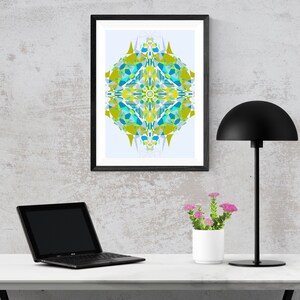 May include: A framed abstract geometric print with a blue, green, and yellow color scheme hangs on a light gray wall above a white desk. A black lamp with a dome-shaped shade sits on the desk next to a potted plant with pink flowers. A black laptop computer is also on the desk.