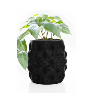 May include: A black, textured plant pot with a wavy design, filled with soil and a green leafy plant. The pot has a unique, modern aesthetic, suitable for indoor or outdoor use. The plant has large, heart-shaped leaves.