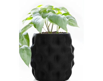 May include: A black plant pot with a textured, wavy pattern, containing soil and a green leafy plant. The pot's design is modern and distinctive. The plant has large, heart-shaped leaves. The pot is suitable for indoor or outdoor use.