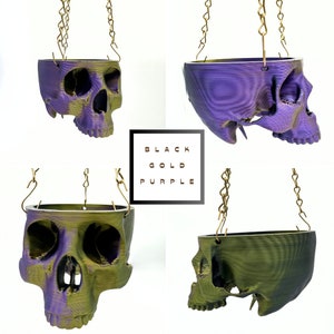 May include: Four hanging skull planters in black, gold, and purple. The planters are made of 3D printed plastic and have a textured surface. The chains are gold.