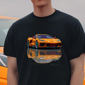 May include: Black t-shirt featuring a vibrant orange sports car graphic. The car is depicted with detailed headlights and a sleek design, reflecting on a glossy surface. The shirt is worn by a person standing in front of a matching orange car.