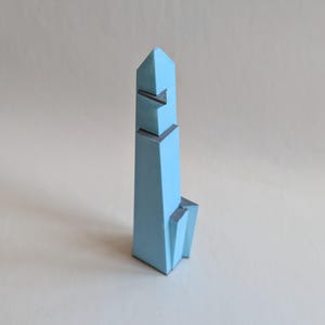 Può includere: Una scultura a forma di torre di colore azzurro, caratterizzata da molteplici sfaccettature con bordi geometrici netti e angolari. L'oggetto presenta una colonna centrale verticale alta con sporgenze a gradini sui lati e una sommità appuntita.