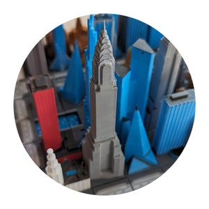 May include: A 3D printed model of a cityscape with a gray skyscraper in the center, surrounded by blue and red buildings.