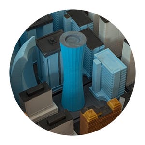 May include: A miniature cityscape model featuring various buildings in shades of blue, gray, and brown. A tall, cylindrical blue structure stands prominently among the other buildings. The model is set against a dark gray base.