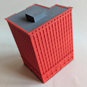 Peut inclure: Un modèle réduit de bâtiment rouge avec un toit gris et une petite structure rectangulaire sur le dessus. Le bâtiment a une façade texturée avec des lignes verticales et une base détaillée. Le modèle est incliné, mettant en valeur son design architectural.