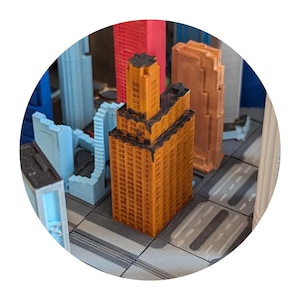 Peut inclure: Un bâtiment miniature orange avec des bordures noires et de nombreuses fenêtres. Le bâtiment fait partie d'une maquette de ville avec d'autres bâtiments en bleu, rouge et marron.