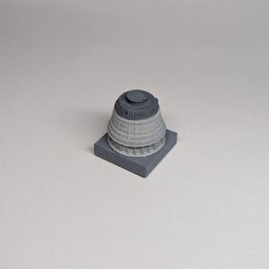 May include: A grey architectural model of a building, possibly a capitol building or similar structure. The model has a cylindrical top and a rectangular base, all in shades of grey.