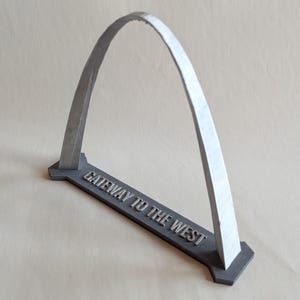May include: A silver-colored arch sculpture with a gray base. The base has the text "GATEWAY TO THE WEST" in raised lettering. The arch is a prominent, curved structure, and the sculpture is set against a neutral background.