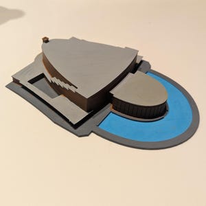以下が含まれることがあります： 曲線的なデザインと青いプールエリアを備えた灰色の建築模型。モデルは、濃い灰色のベースと明るい灰色のメイン構造を持つ、層状の構造をしています。全体的なデザインはモダンで幾何学的です。