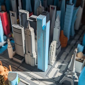 May include: A 3D printed miniature city model featuring a central light blue skyscraper surrounded by various grey, white, and orange buildings. The layout includes detailed architectural structures like the Chrysler Building and an Eiffel Tower replica.