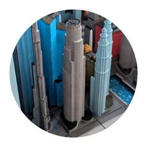 May include: A close-up of a 3D printed model of a city skyline. The buildings are made of gray, blue, and white plastic. The model is detailed and realistic.