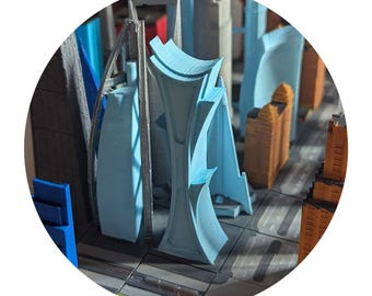 May include: A close-up of a 3D printed model of a city skyline. The model features several buildings, including a tall, blue, modern skyscraper with a unique design.