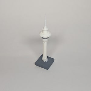May include: A small, white model of a tower with a spire, mounted on a grey square base. The tower features a unique design with a rounded top and a thin, cylindrical structure. This model is likely a souvenir or architectural replica.