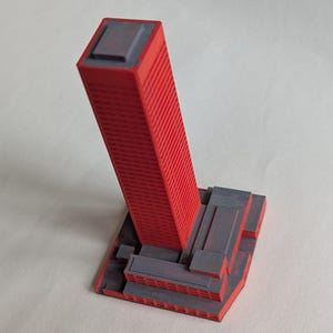 May include: A 3D-printed model of a skyscraper. The building is primarily red with a gray top and base. The tall, rectangular structure has vertical lines, and the base features a stepped design.