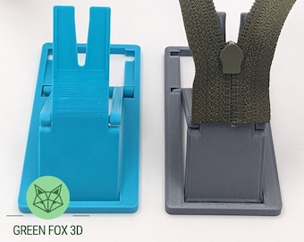 May include: A 3D printed foldable zipper jig in blue and gray. The jig is designed to hold a zipper in place while sewing. The jig is folded up and the text "GREEN FOX 3D" is visible.