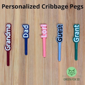 May include: Five personalized cribbage pegs in maroon, blue, pink, light blue, and green. Each peg features a vertical name or title in white lettering: Grandma, Dad, Lori, Guest, and Grant. The pegs have a slender cylindrical base.