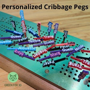 May include: A collection of personalized cribbage pegs in various colors, including blue, red, purple, and black. Each peg features a name, with the text "Personalized Cribbage Pegs" at the top. The pegs are arranged on a green metal cribbage board.