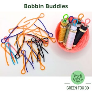 May include: A collection of colourful bobbin buddies, small plastic loops used to hold thread spools, are scattered on a white surface. A bowl filled with spools of thread is in the background. The bowl is pink and has a textured surface. The text "GREEN FOX 3D" is at the bottom of the image.