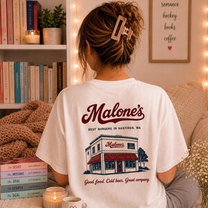 Puede incluir: Una camiseta blanca con un gráfico en la espalda. El diseño muestra un restaurante con un toldo rojo, el texto "Malone's", "BEST BURGERS IN HASTINGS, MA", "EST. 1942" y la frase "Good food. Cold beer. Great company." en colores rojo y azul.