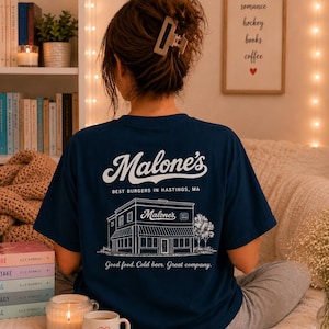 May include: A navy blue t-shirt featuring a white graphic on the back. The design includes the text "Malone's", "BEST BURGERS IN HASTINGS, MA", and "Good food. Cold beer. Great company." along with a line art illustration of a restaurant building.