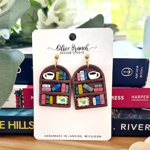 Peut inclure: Une paire de boucles d'oreilles pendantes marron en forme d'arche représentant une bibliothèque miniature. Chaque boucle présente des livres colorés, une tasse à café blanche et un petit cadre. Elles sont fixées sur une carte Olive Branch Design Studio.