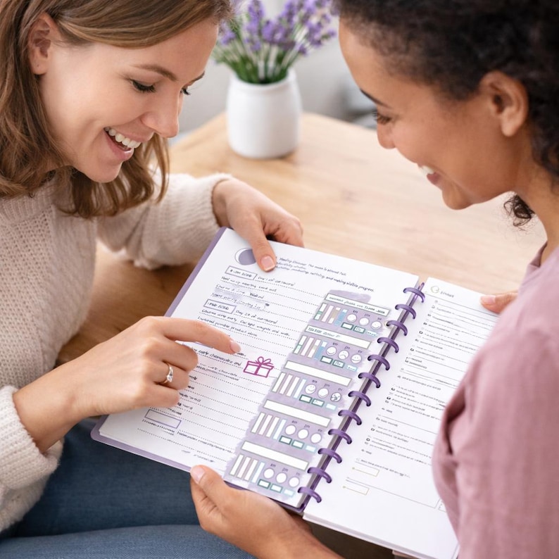May include: Two people are looking at a planner with a purple spiral binding. The planner pages have a white background with purple and grey accents. The planner is open, and one person is pointing at a section with a gift icon.