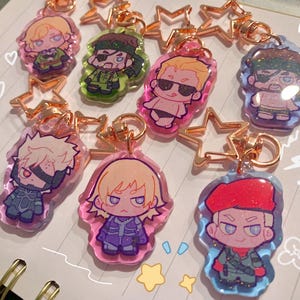 May include: A selection of acrylic keyrings featuring cartoon characters. Each keyring has a different character design, with gold-coloured star-shaped clasps. The keyrings are in various colours, including pink, green, and blue.
